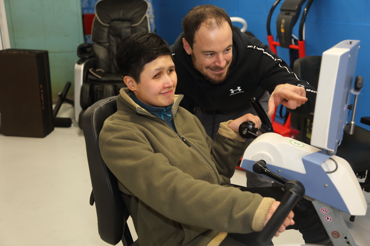 Matt Brinkley providing hands-on support and guidance to a member using accessible exercise equipment in the therapy gym, demonstrating the personalized approach at The Angel Centre
